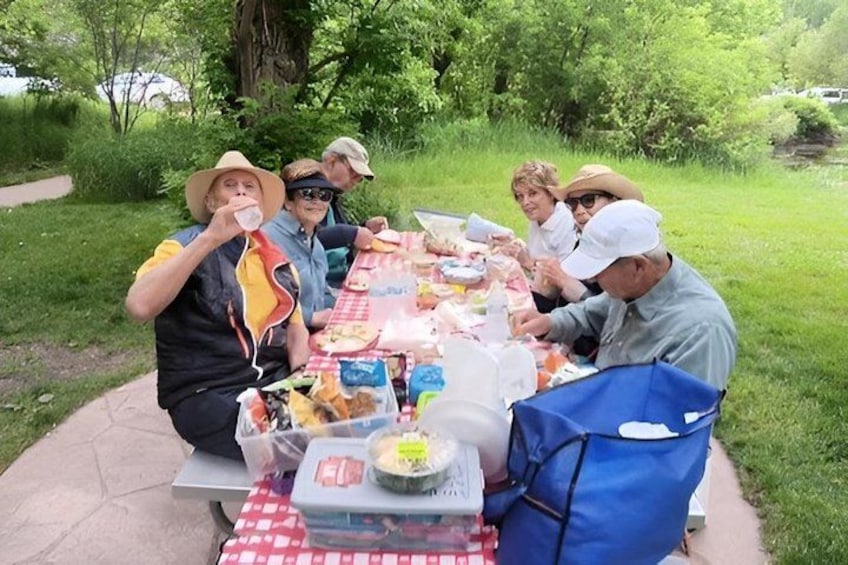 A group of people sitting at an outdoor picnic table with food and drinks. 2