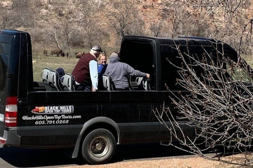 A black van with a sign for Black Hills Open Top Tours parked in a natural setting with trees and a clear sky. 1