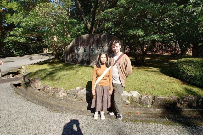 Two people standing in front of a large stone monument with Japanese characters, surrounded by trees and a gravel path. 4