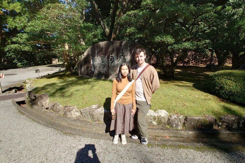 Two people standing in front of a stone monument with Japanese characters, a paved path, and a grassy area with trees. 2