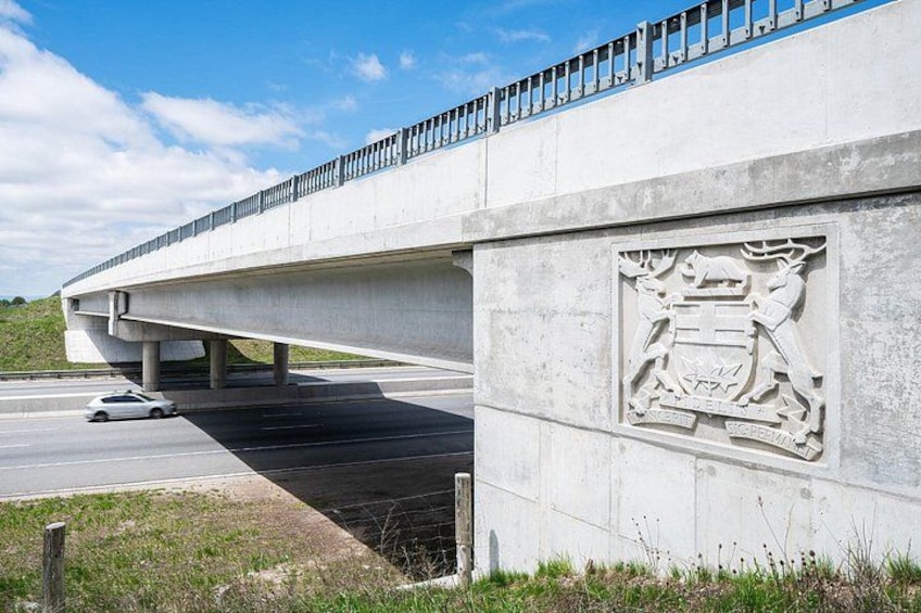 Eine Betonbrücke, unter der ein Auto fährt, mit einem dekorativen Wappen an der Seite. 3