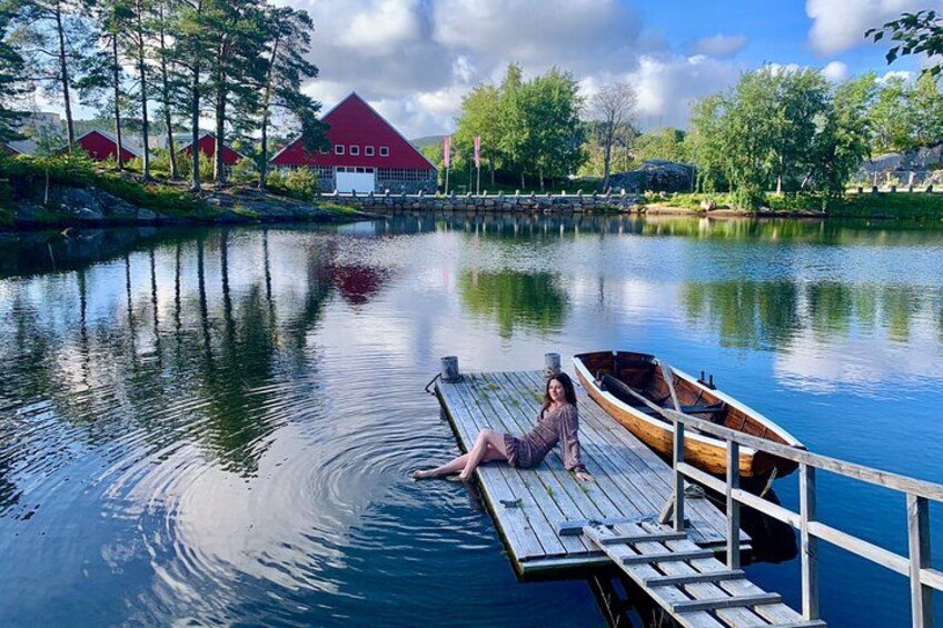Une femme est assise sur un quai, au bord d’un lac paisible. Un bateau en bois est amarré à proximité, et on aperçoit des bâtiments rouges en arrière-plan. 1