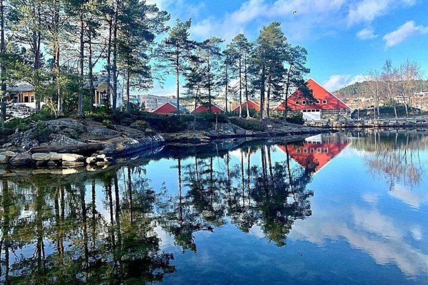 Un lago tranquilo con reflejos de árboles y edificios, rodeado de un bosque y algunas casas con tejados rojos. 4