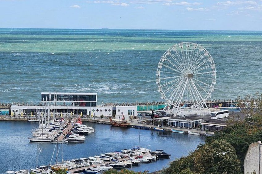 A coastal marina with a large Ferris wheel, boats, and a building with a modern design. 3