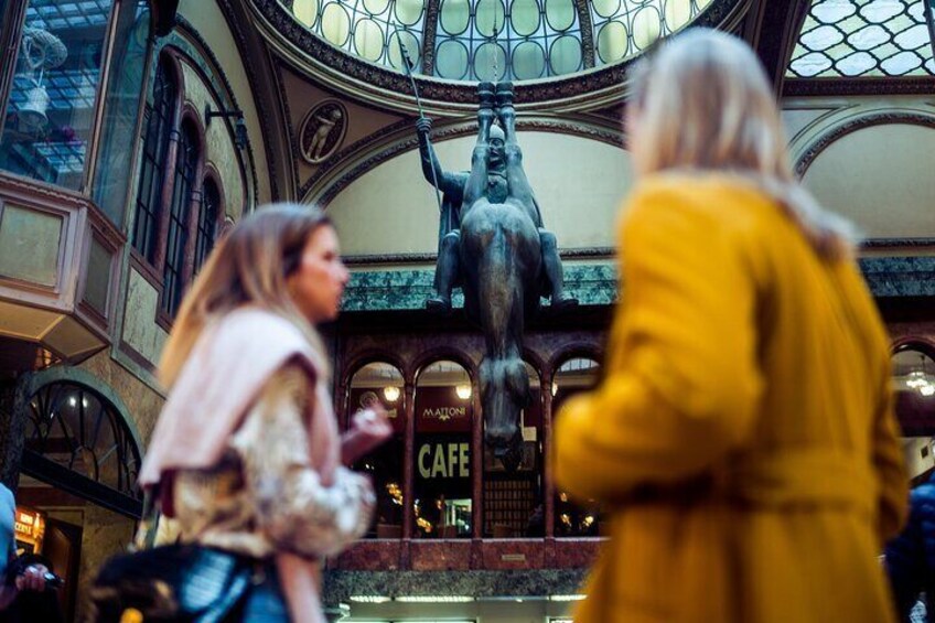 Una mujer con abrigo amarillo camina frente a una estatua en un edificio de ventanales arqueados y cartel de café. 5