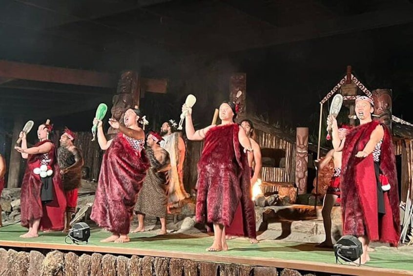 A group of people performing a traditional dance on a stage with a rustic backdrop. 2