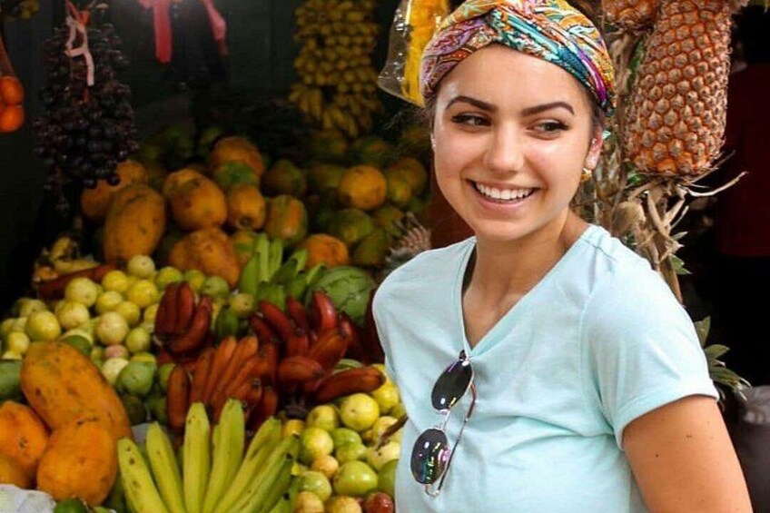 A person standing in front of a fruit market with various fruits like bananas, pineapples, and oranges. 1