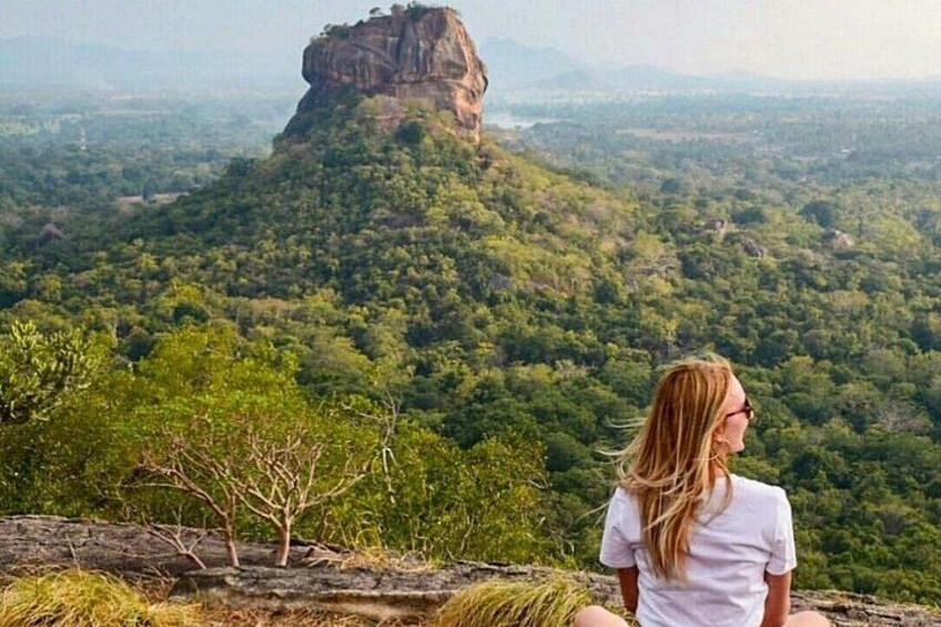 A person sitting on a rock overlooking a forested landscape with a prominent rock formation in the distance. 3