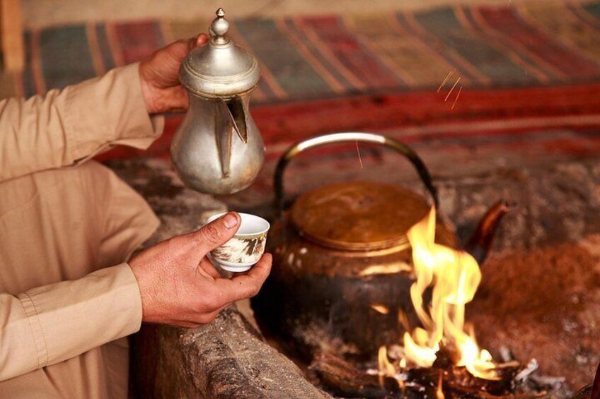 A person pouring tea from a metal pot into a cup, with a traditional teapot and a fire burning in the background. 3