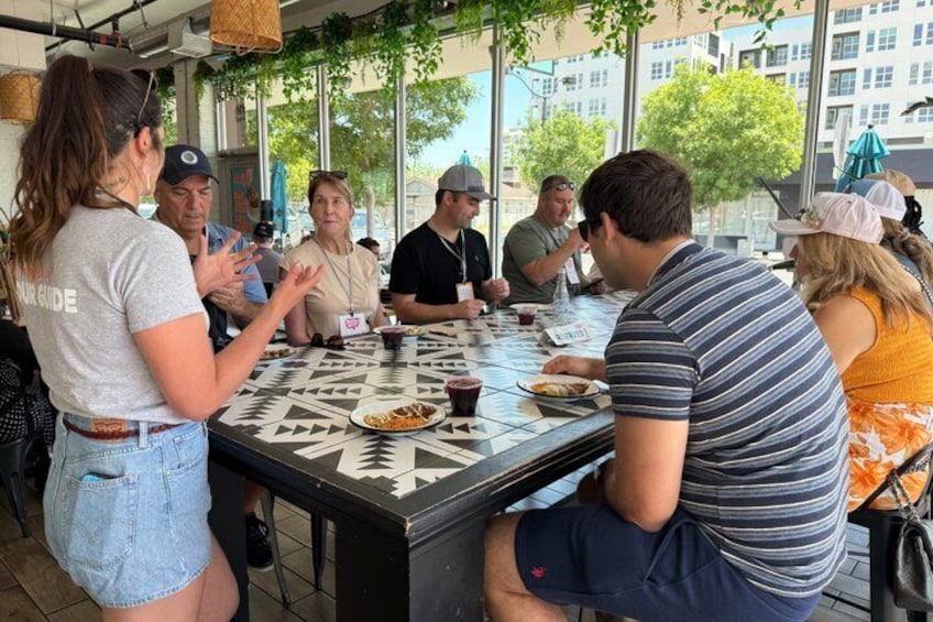 A group of people seated around a table with food and drinks, outdoors with trees and buildings in the background. 1
