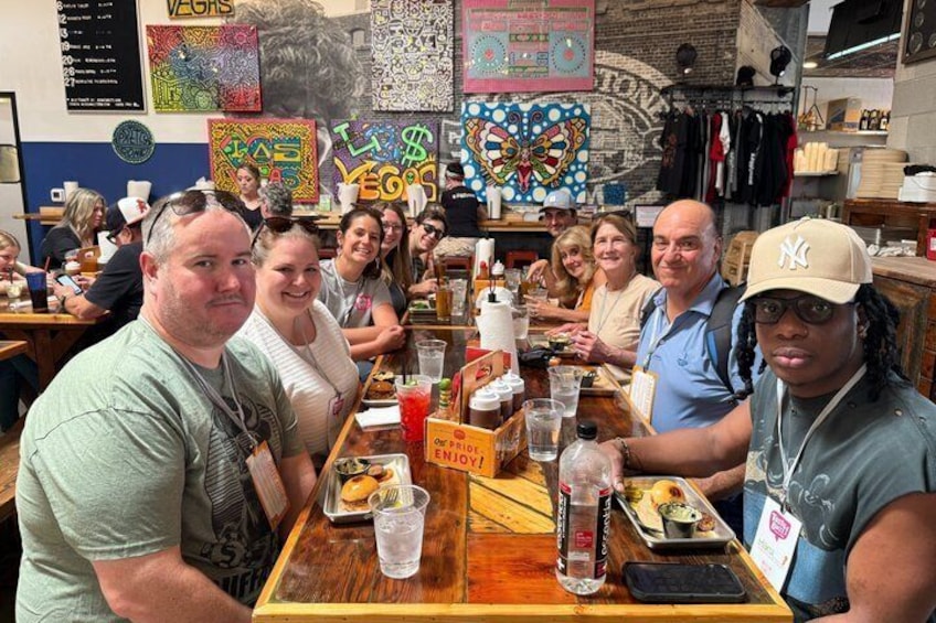 A group of people seated at a wooden table in a restaurant with colorful artwork on the walls. 3