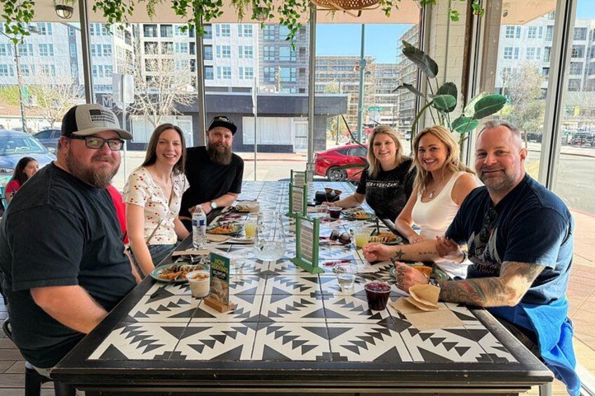 A group of people dining at an outdoor restaurant with a patterned tablecloth and a view of buildings in the background. 4