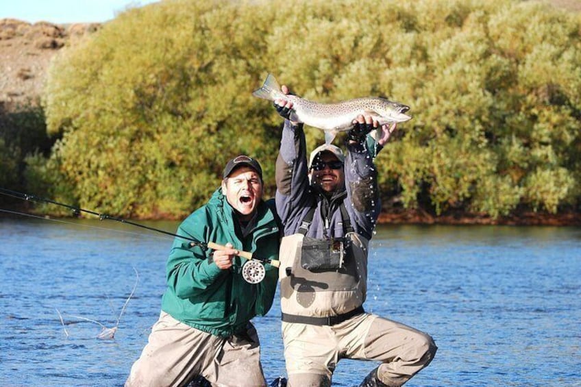 Two people holding a fish, standing in a river with trees in the background. 1