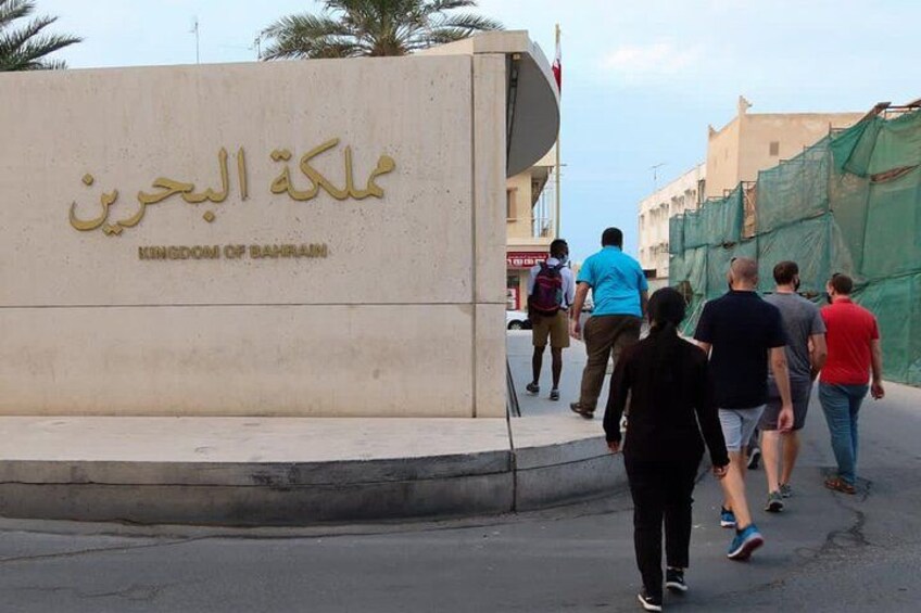 A group of people walking past a sign that reads "Kingdom of Bahrain" in Arabic. 1