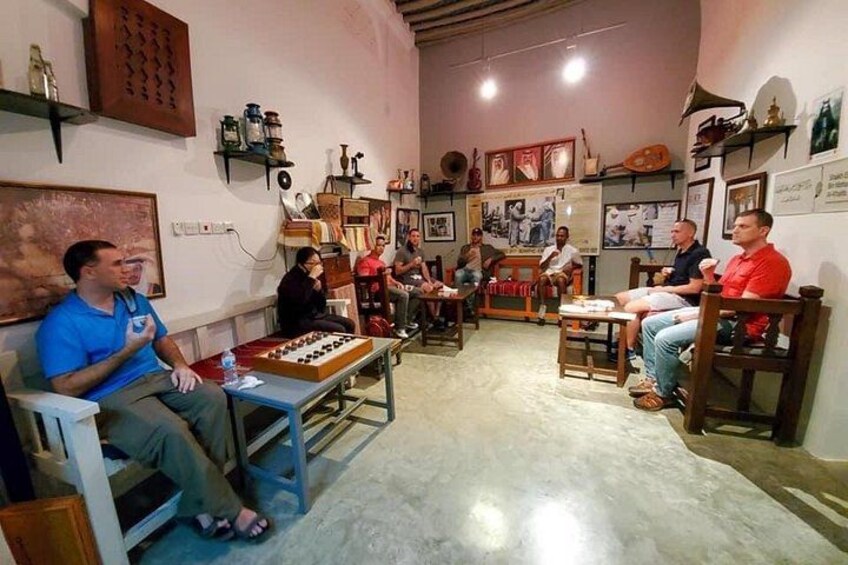 A group of people seated in a room with various items on the walls and a chessboard on the table. 2