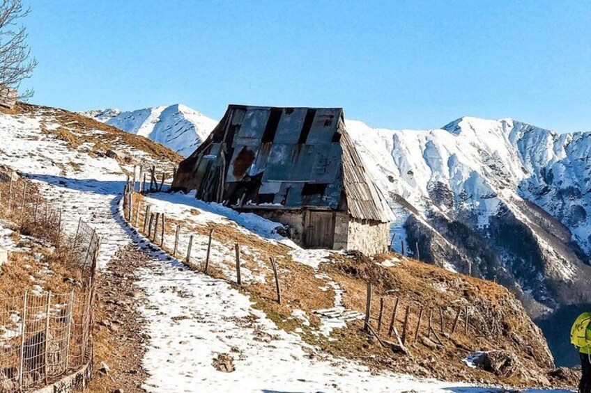 Sendero nevado que asciende hacia una estructura de madera en ruinas con techo de metal, rodeado de picos nevados. 2