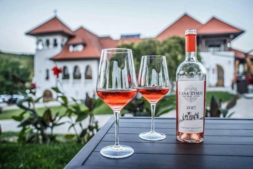 Una botella de vino Casa Timis 2017 y dos copas de vino tinto sobre una mesa, con un edificio al fondo. 5