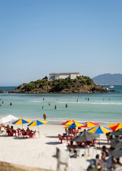 Une plage où l’on voit des gens nager et bronzer, des parasols colorés, et un bâtiment sur une petite île en arrière-plan. 3