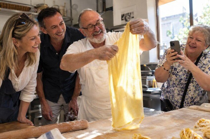 A group of people in a kitchen, one person holding a yellow bag, another taking a photo, and pasta on the counter. 1