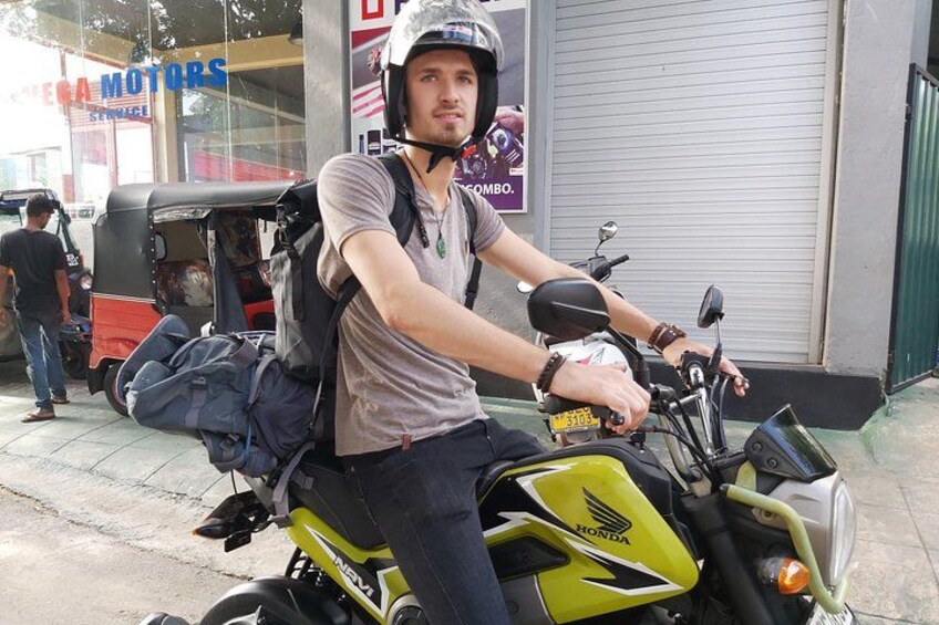 A person on a motorcycle with a backpack, parked in front of a building with a sign that reads 'FEMA MOTORS'. 1
