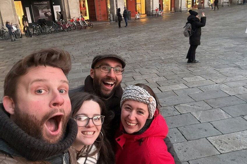Un gruppo di persone si scatta un selfie in una piazza con pavimentazione in pietra, mentre una persona sullo sfondo scatta una foto. 4