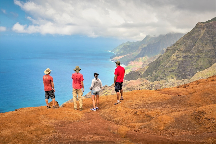 Four people standing on a reddish-brown hillside overlooking a blue ocean and green mountains. 1