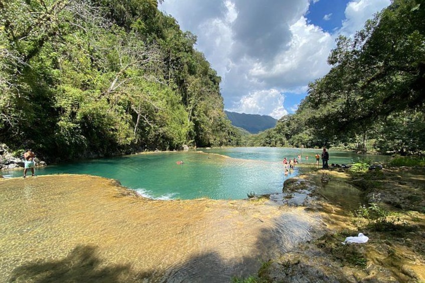 A turquoise river surrounded by dense forest, with people swimming and standing on the banks. 5