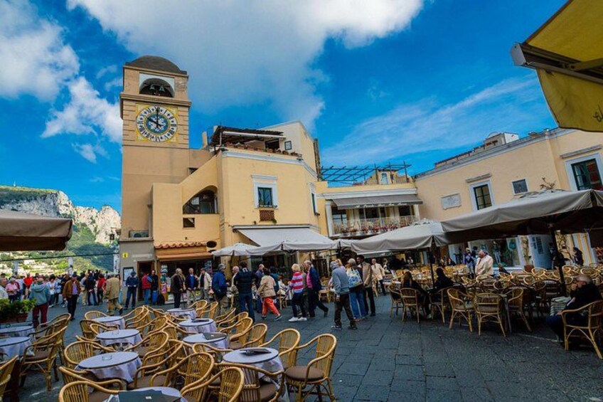 A bustling outdoor café area with wicker tables and chairs, a clock tower, and a mountainous backdrop. 2
