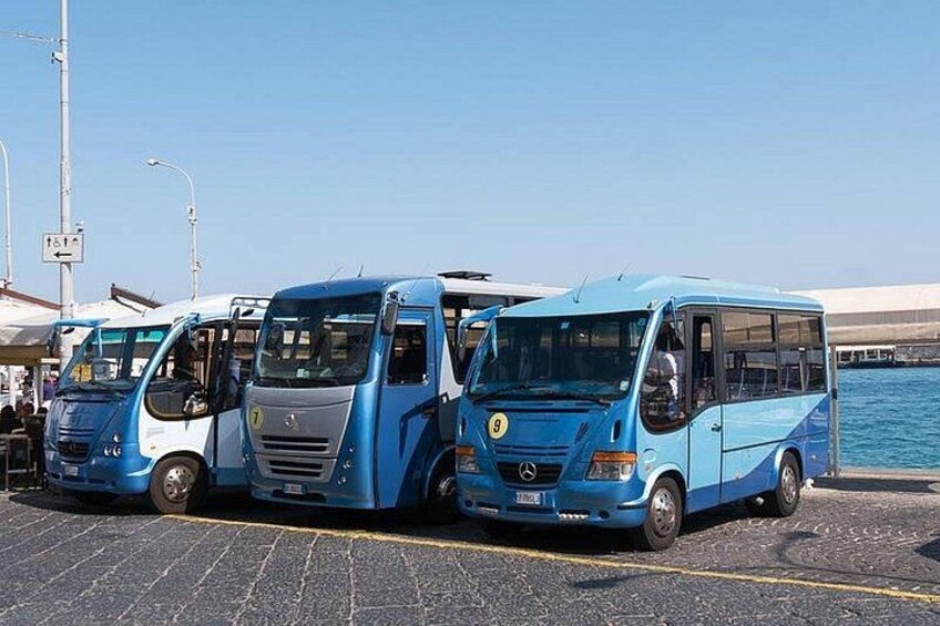 A row of blue buses parked in a lot by a waterfront. 3