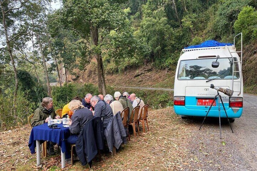 A group of people dining outdoors at a table set up on a gravel path with a van parked nearby. 1