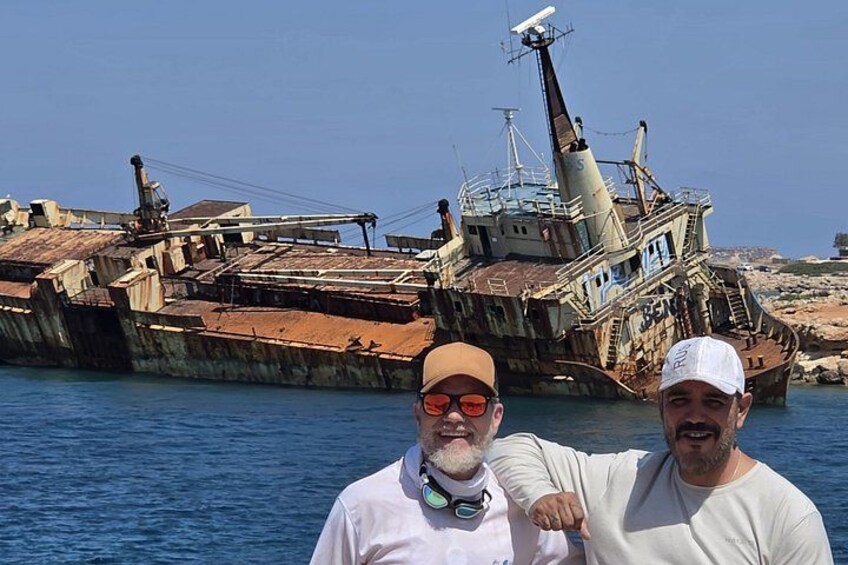 Two men standing in front of a rusted shipwreck in a body of water. 1