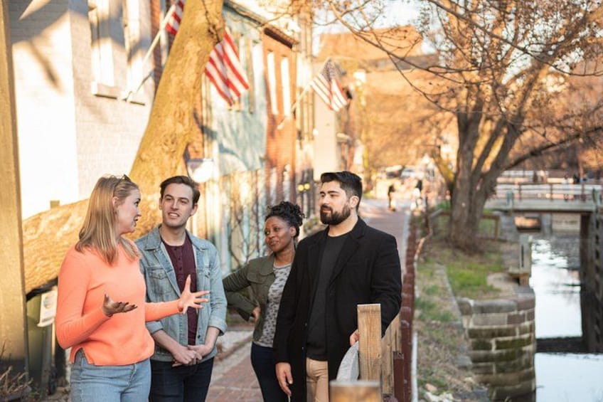 A group of people standing on a sidewalk next to a canal with buildings and an American flag in the background. 1