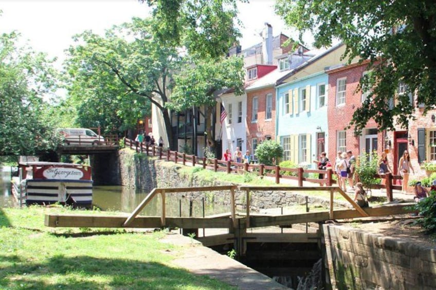Un puente de madera sobre un canal, con personas caminando, rodeado de edificios coloridos y vegetación. 2