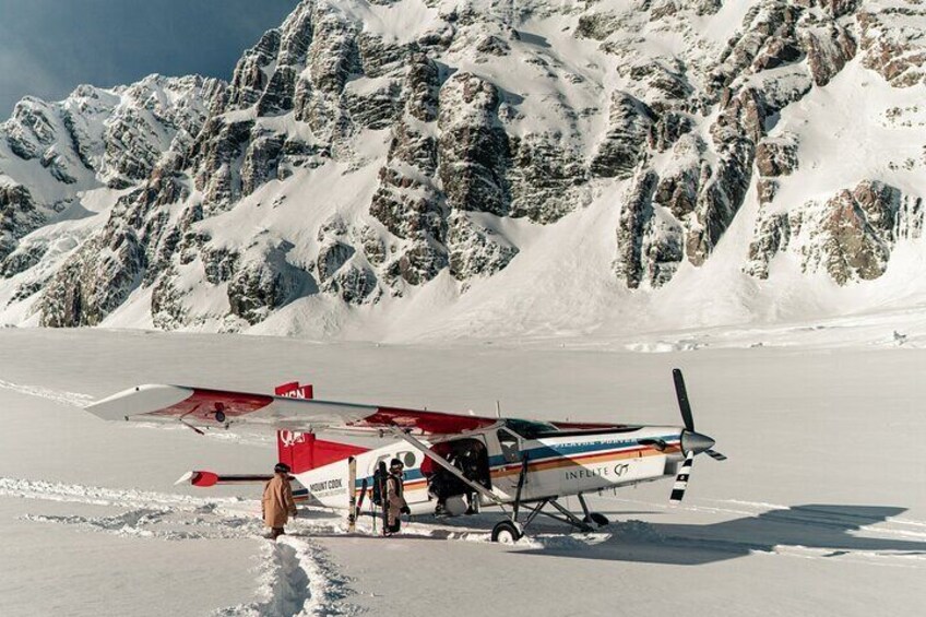 A small aircraft with a red and white livery parked on snow-covered ground with mountains in the background. 5