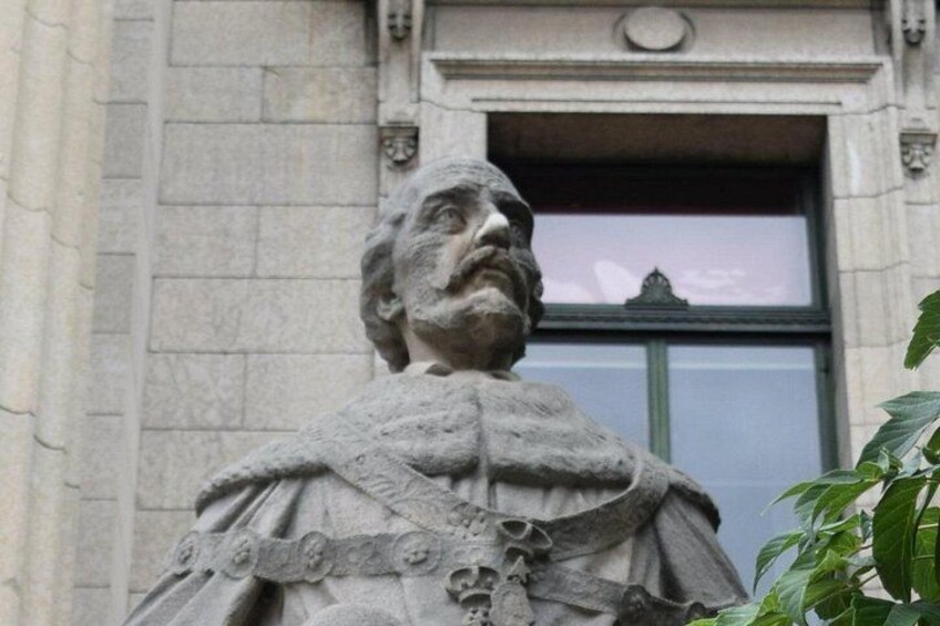 A statue of a man with a beard, wearing a robe with a cross, in front of a building with a window and a stone wall. 3