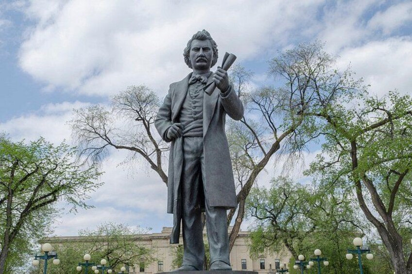 A statue of a man in a long coat holding books, set against a backdrop of trees and buildings. 2