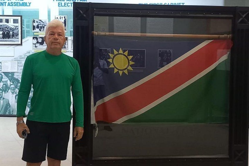 A man standing next to a framed flag with a sun emblem, in a room with a glass display case and informational posters. 4