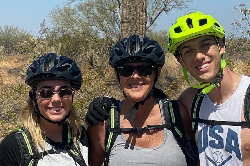 Three people wearing helmets and cycling gear, standing outdoors with trees and shrubs in the background. 2
