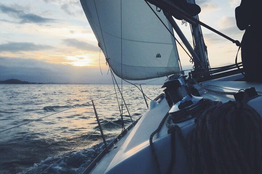 A sailboat with a white sail and ropes on the deck, sailing on a body of water with a sunset in the background. 1