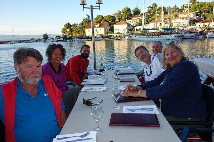 A group of people sitting at a table by a waterfront, with boats and buildings in the background. 3