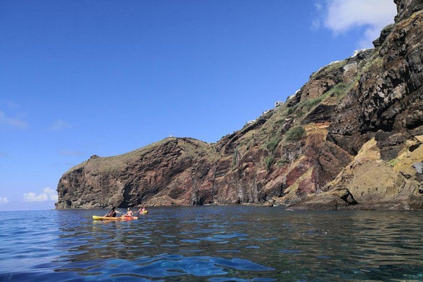 A rocky coastline with a clear blue sky, calm sea, and a person kayaking. 2