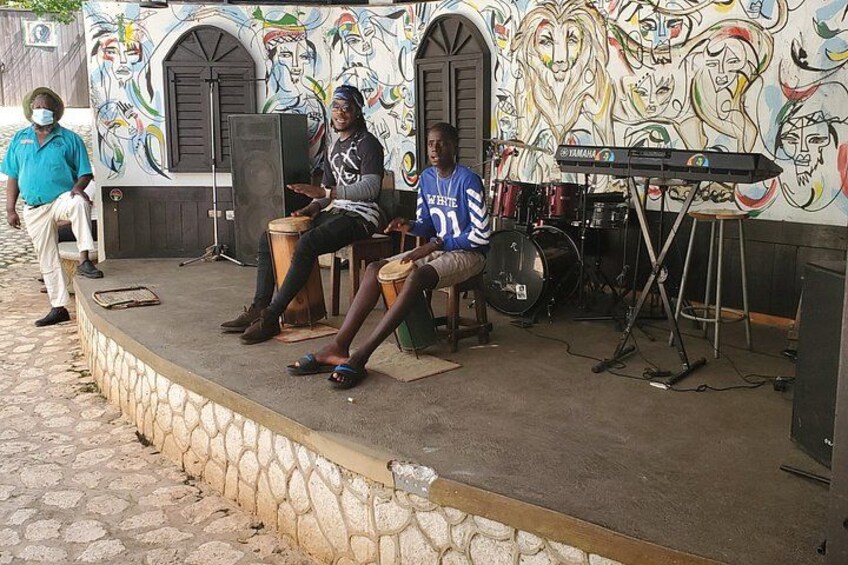 A street musician performing with drums and a keyboard, seated on a stage with a mural in the background. 3