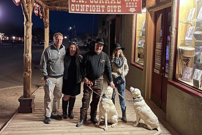 A group of people and dogs on a wooden deck with a sign reading 'C.R. CORRAL GUNFIGHT SITE' above. 2
