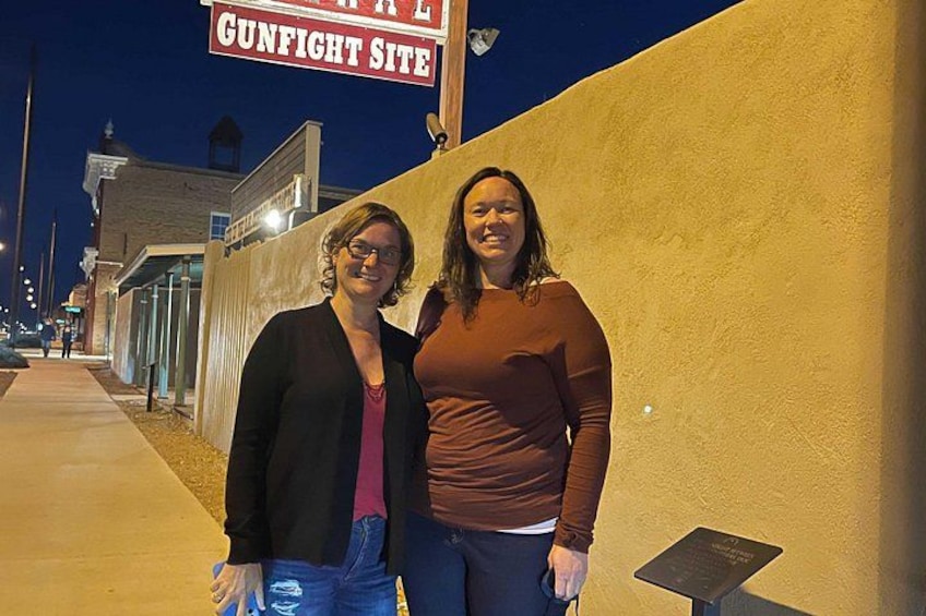 Two people standing in front of a building with a sign that reads 'GUNFIGHT SITE.' 5