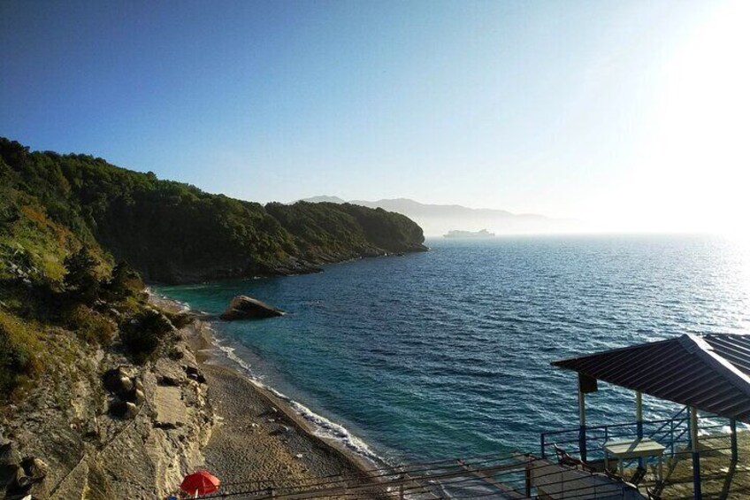 A coastal landscape with a rocky beach, a clear blue sea, and a forested hillside. 1