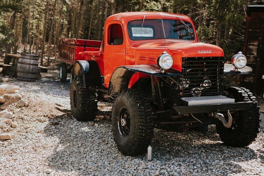 A red Dodge truck with off-road tires and a bed, parked on a gravel path in a wooded area. 5