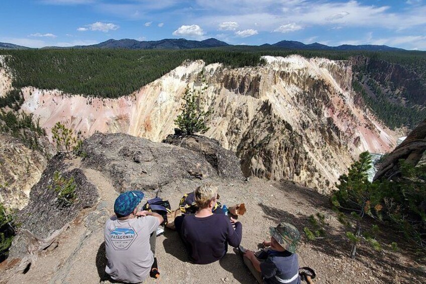 A group of people sitting on a rocky outcrop overlooking a canyon with distinct layers of rock and a forested valley in the background. 2