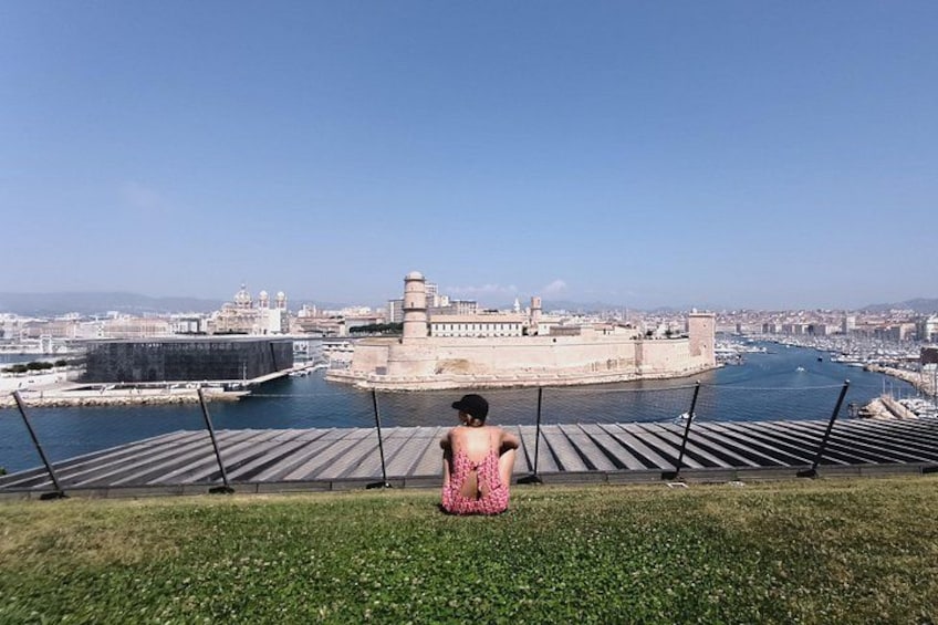 Una persona sentada en una cubierta de madera contemplando una ciudad amurallada con un puerto. 3