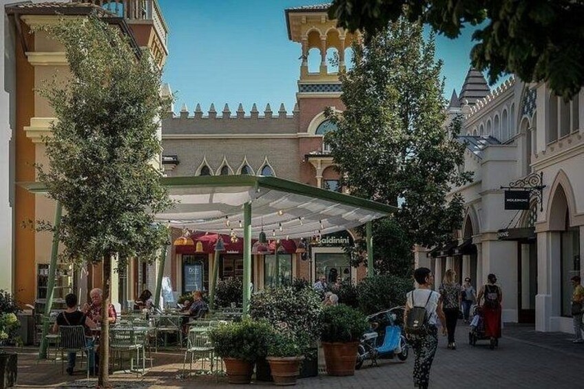 A covered outdoor café with tables and chairs, surrounded by buildings with arched windows and a castle-like structure in the background. 2