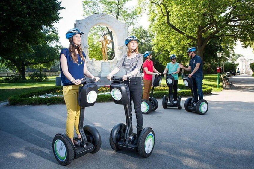 Mensen rijden op segways op een verhard pad in een park, met bomen en een stenen boog in de achtergrond. 5